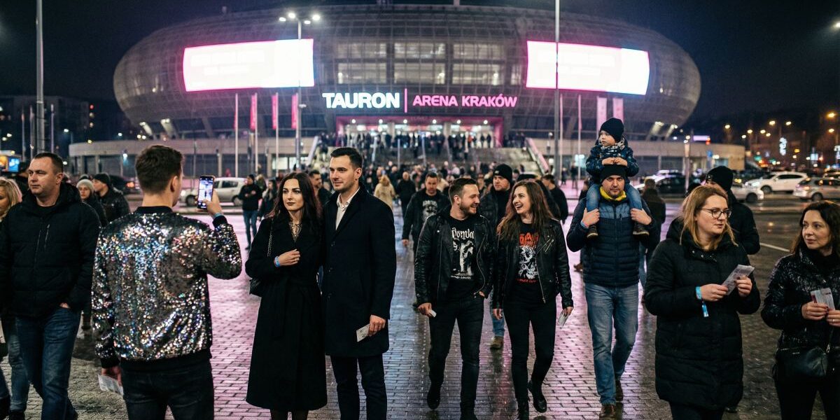 Nocna scena przed Tauron Areną Kraków, z jasno podświetlonym napisem i tłumem uśmiechniętych ludzi wychodzących po wydarzeniu – pary, grupy znajomych i rodziny z dziećmi, co oddaje atmosferę miasta, które w 2026 roku gra na pełny stadion emocji.